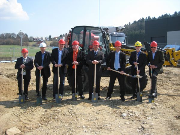 Acht Männer in Anzügen und mit bunten Baustellenhelmen und Schaufeln, vor einem kleinen Bagger.